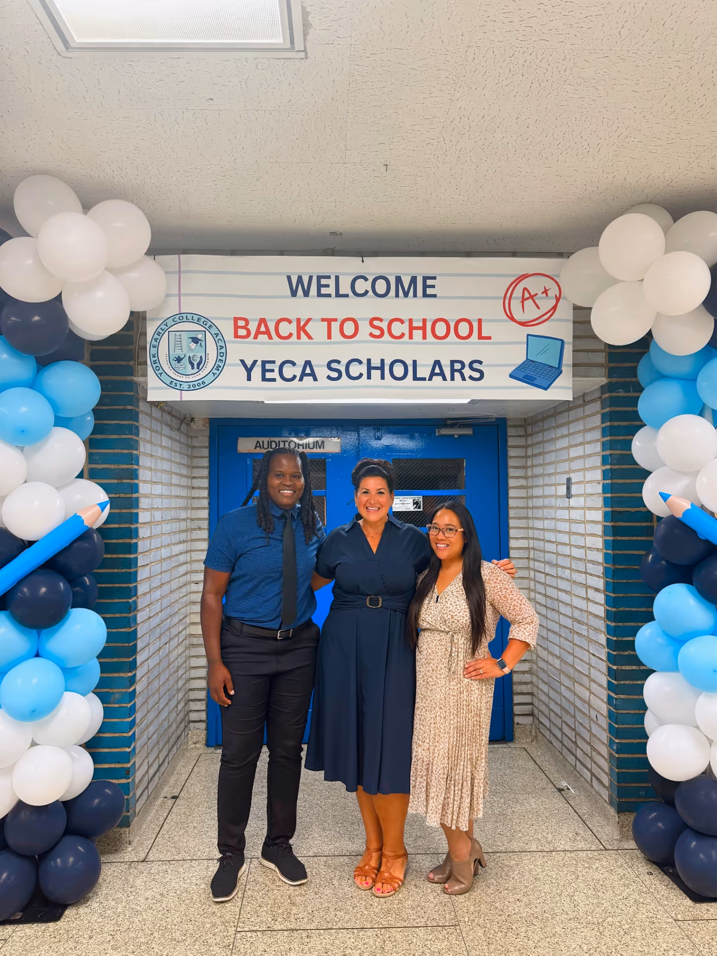 Three people posing under a decorated archway with balloons and a welcome sign.