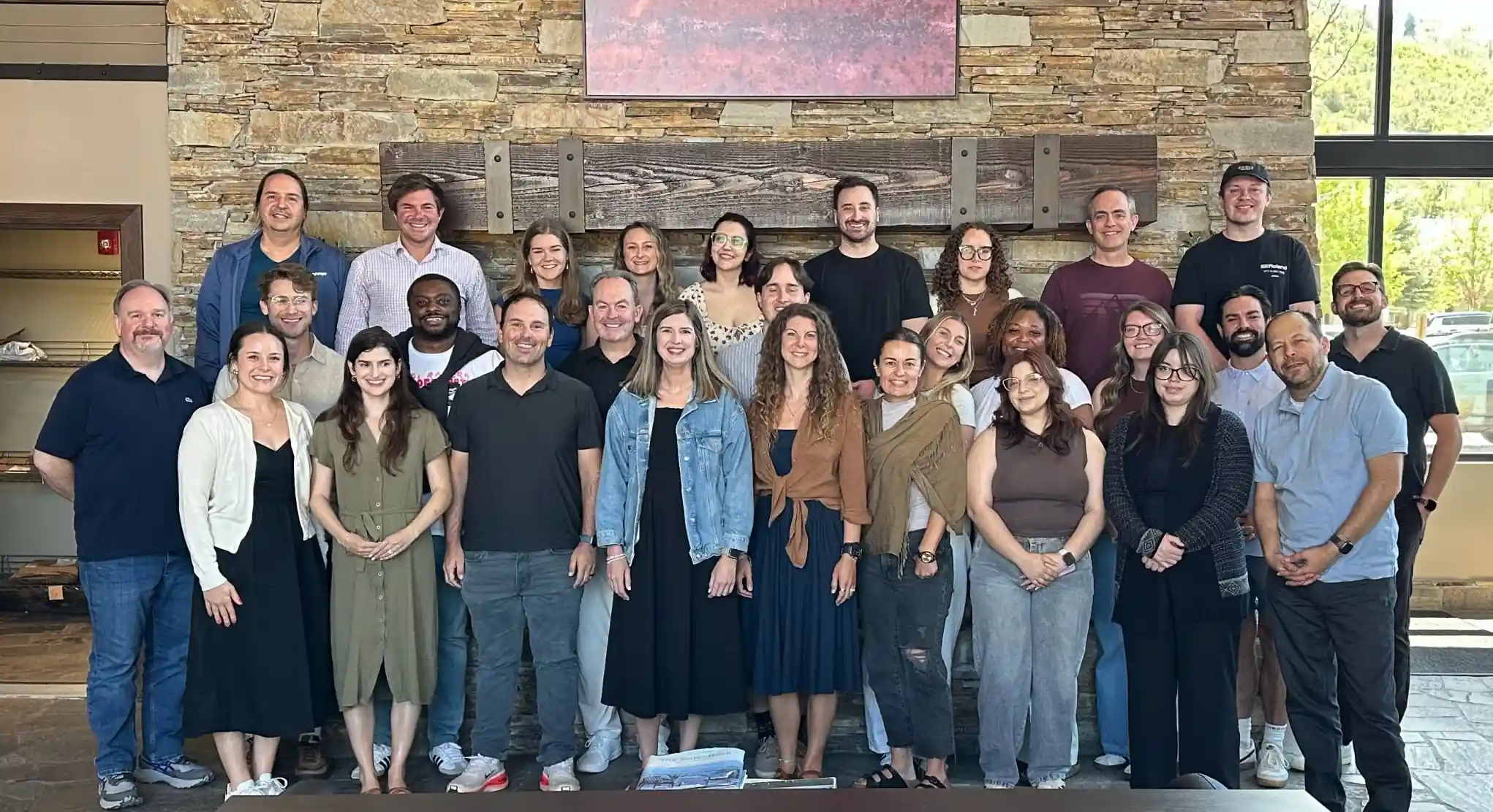 Group photo of 27 diverse people standing and smiling indoors against a stone wall with large windows and wooden decor above.