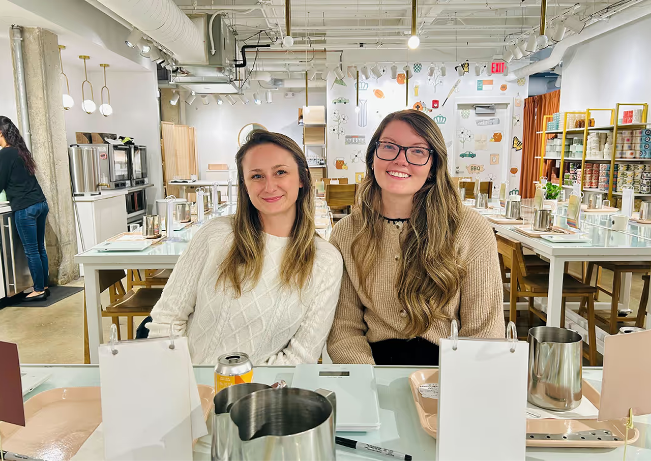Two smiling women sit side by side at a table in a bright room set with metal pitchers, trays, a notebook, and pens, with shelves and a person working in the background.