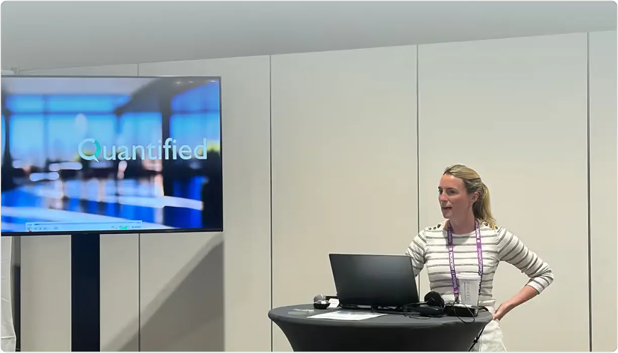 Woman with striped shirt standing behind a round table with a laptop and microphone, presenting next to a screen showing the word 'Quantified' in a modern office setting.