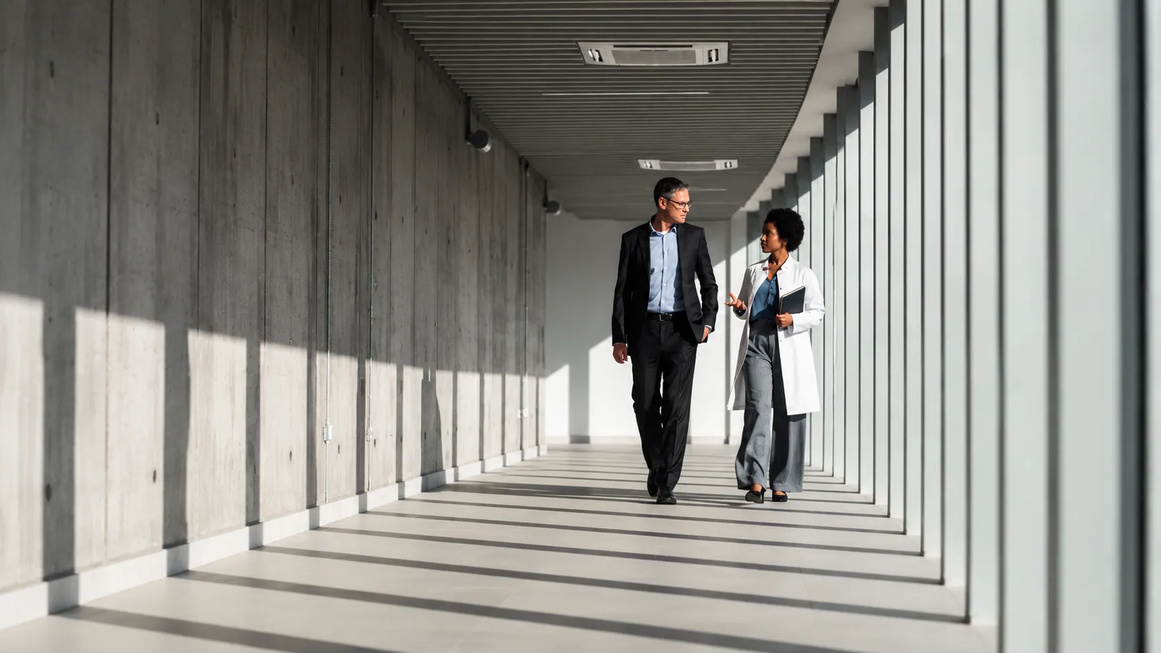 A pharmaceutical sales rep and a physician walking and talking in a sunlit hospital corridor