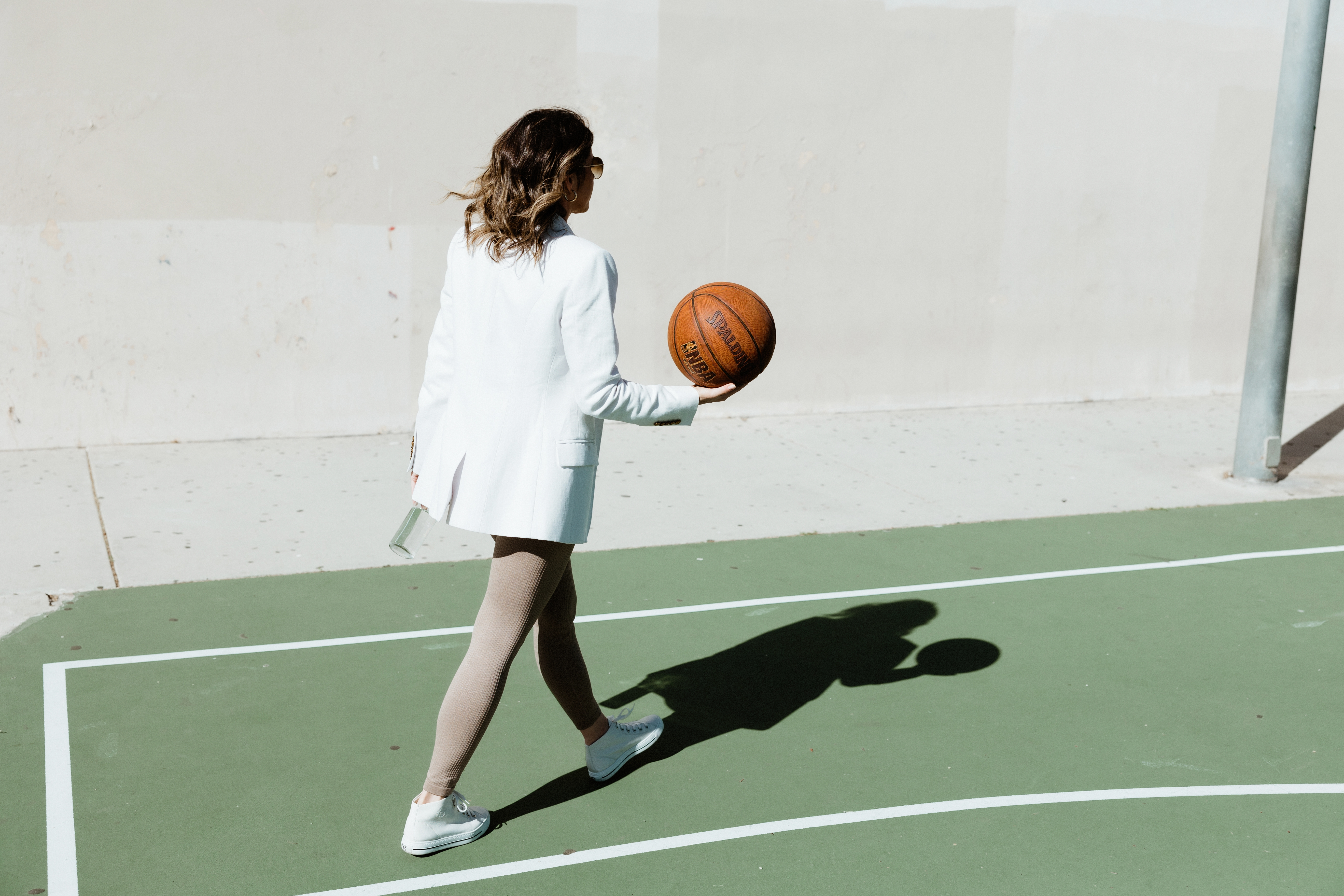 Woman in white blazer and leggings walking on a basketball court holding a basketball and a water bottle.