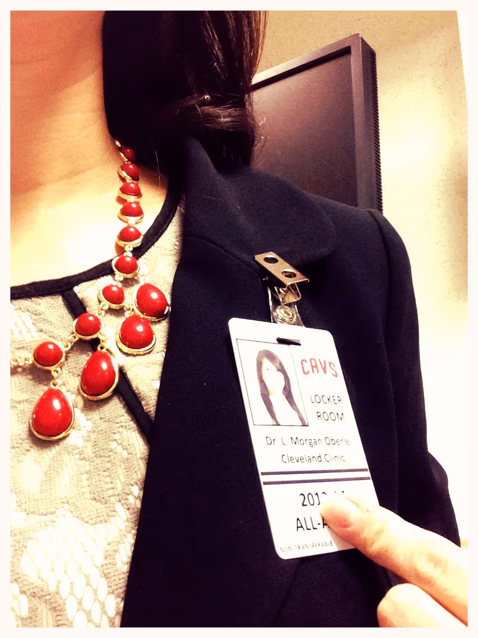 Close-up of a person wearing a black blazer with a Cleveland Clinic locker room ID badge clipped, finger pointing at the badge, and a red beaded necklace visible.
