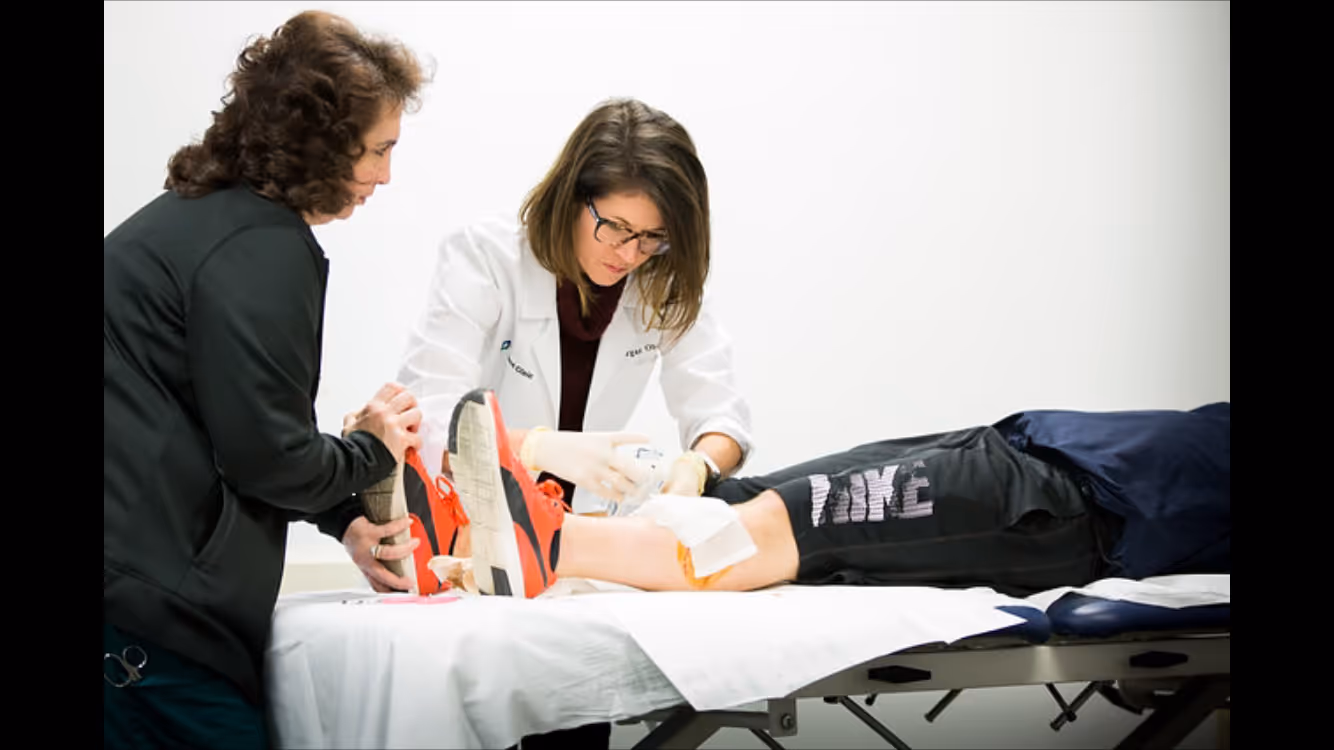 Medical professional in a white coat attending to a patient's injured knee on an examination table while another person supports the patient's foot.