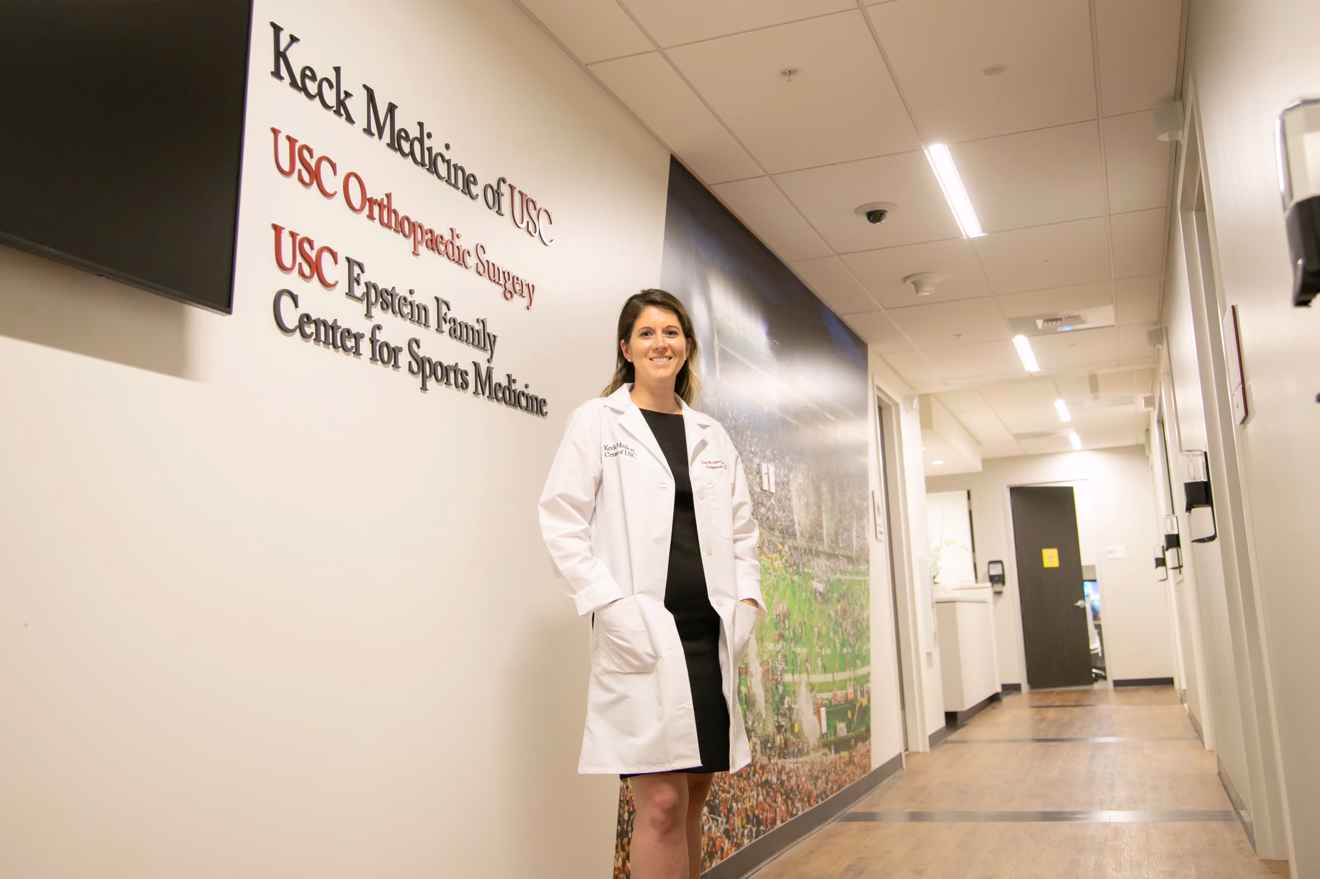 A woman doctor in a white lab coat stands in a bright hallway with Keck Medicine of USC and USC Orthopaedic Surgery signage on the wall.