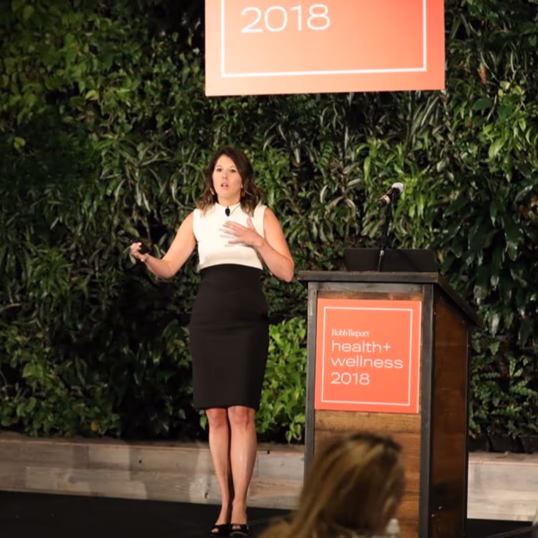 A woman in a black and white dress speaking on stage next to a podium with a sign reading 'Robb Report health + wellness 2018' against a backdrop of green foliage.