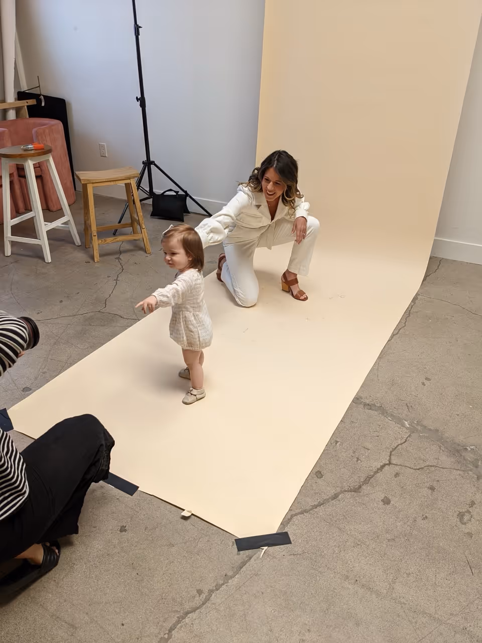 A woman in white clothes kneeling on a beige backdrop holding a toddler who stands and points forward during a photo shoot.