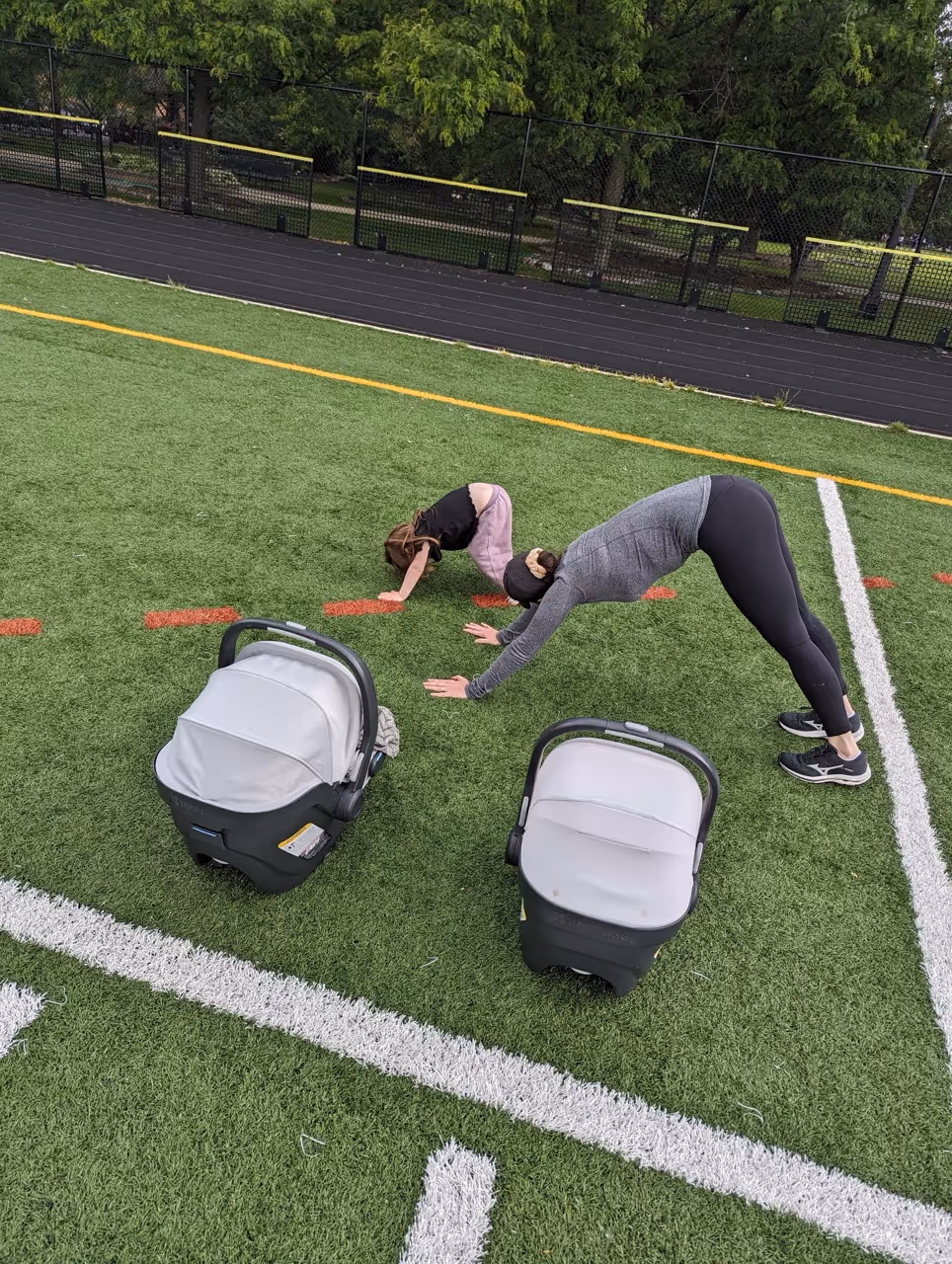 Adult woman and child doing downward dog yoga poses on a green turf field near two covered infant car seats.