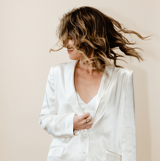 Person with wavy hair wearing a white blazer against a beige background, hair blowing to the side.
