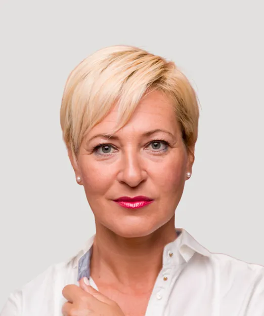 Portrait of a confident middle-aged woman with short blonde hair, wearing a white shirt and pink lipstick.