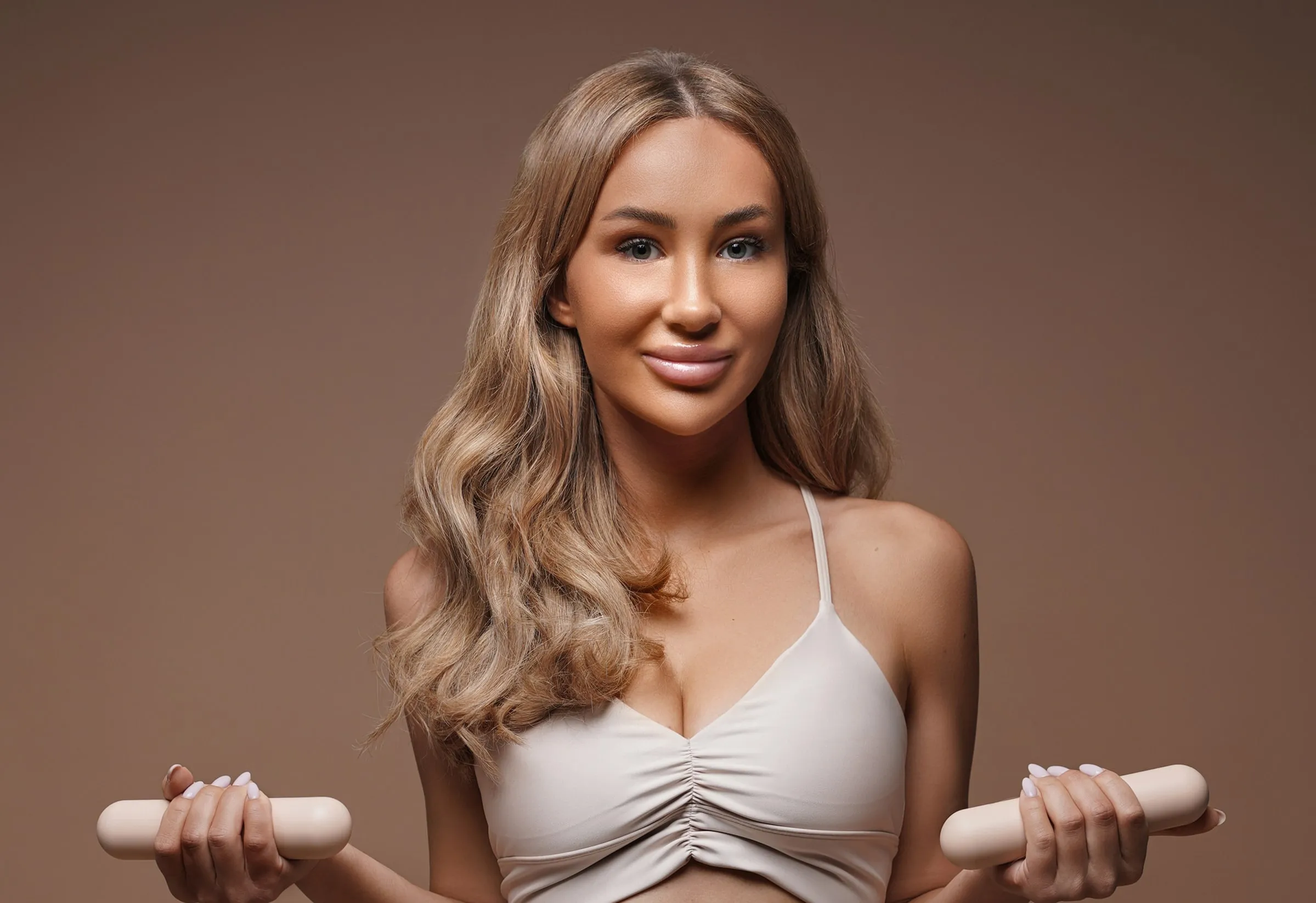 Young woman with long wavy hair wearing a beige sports bra holding beige dumbbells with both hands against a brown background.