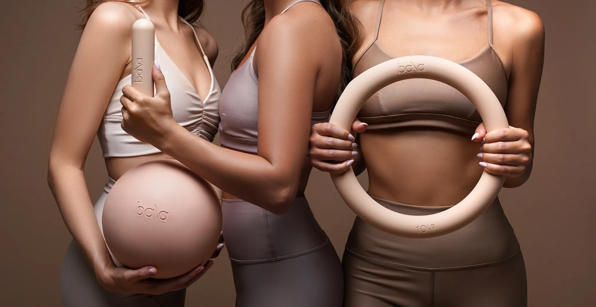 Three women in neutral-toned activewear holding beige Bala fitness weights: a ball, a ring, and a bar.