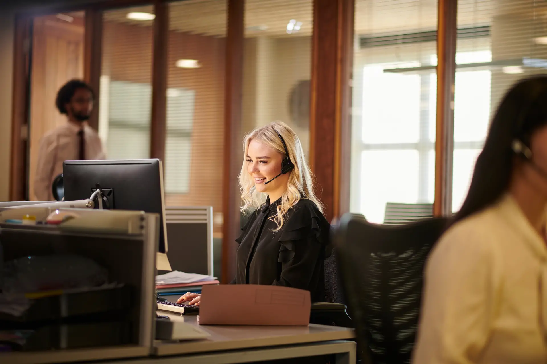 A smiling woman with blonde hair wearing a black blouse and headset, working on a computer in a modern office with wooden walls and glass partitions, with two blurred coworkers in the background.
