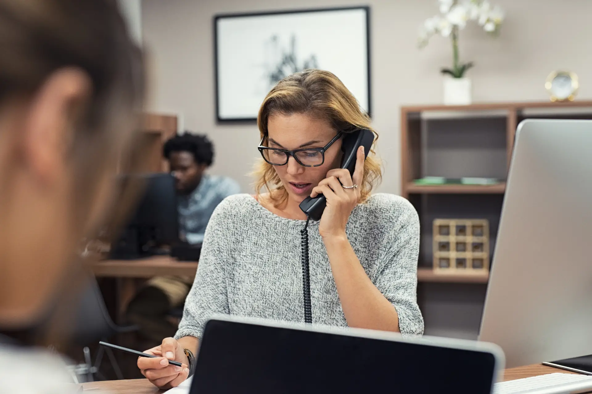 Woman with glasses and a gray sweater speaking on a corded phone while holding a pen, sitting at a desk with a laptop, with a blurred person and office shelves in the background.