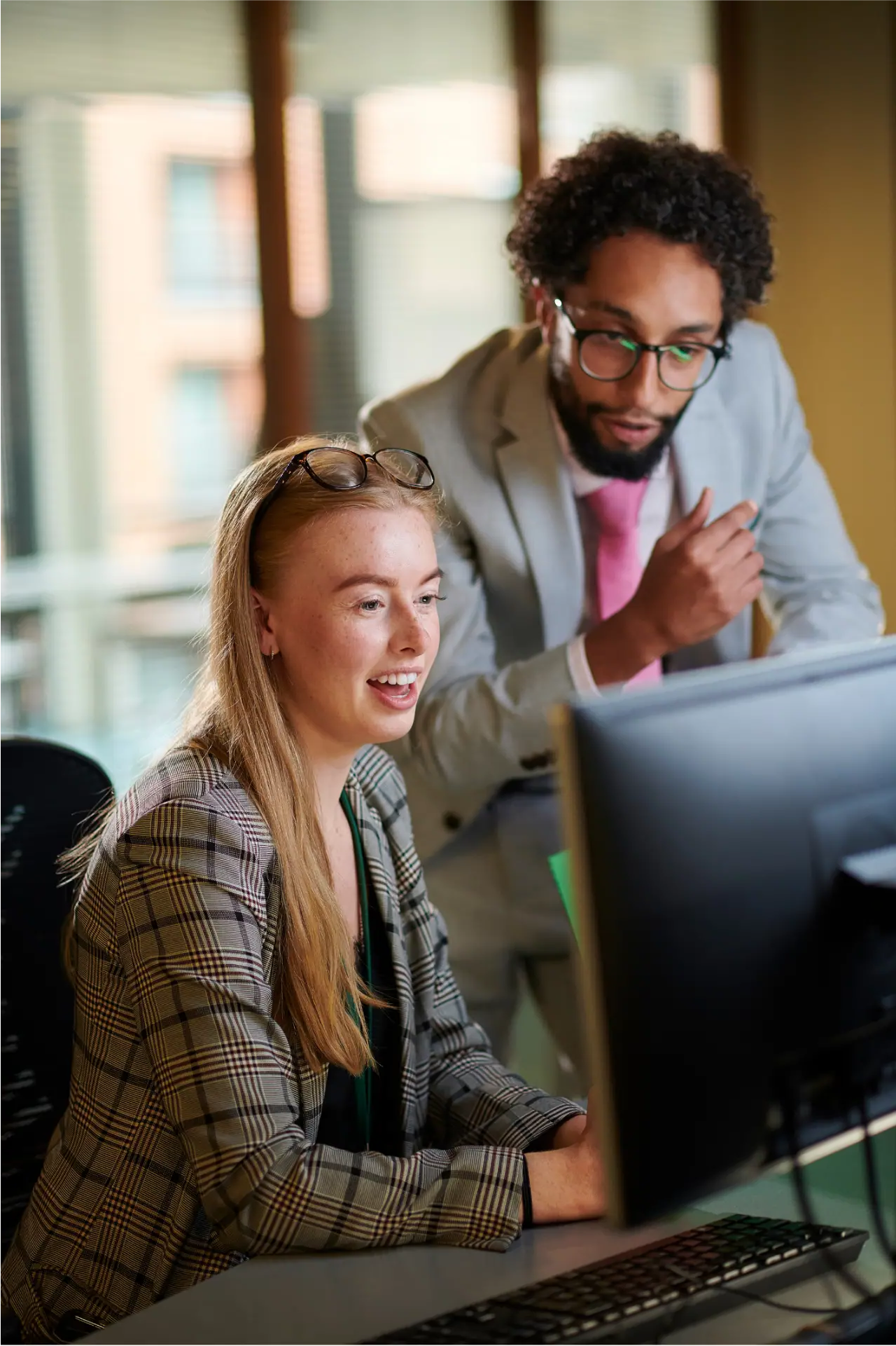 A young woman with long blonde hair wearing a plaid blazer and glasses on her head is smiling and looking at a computer screen while a man with curly hair, glasses, and a gray suit with a pink tie leans over her shoulder pointing at the screen in an office setting.