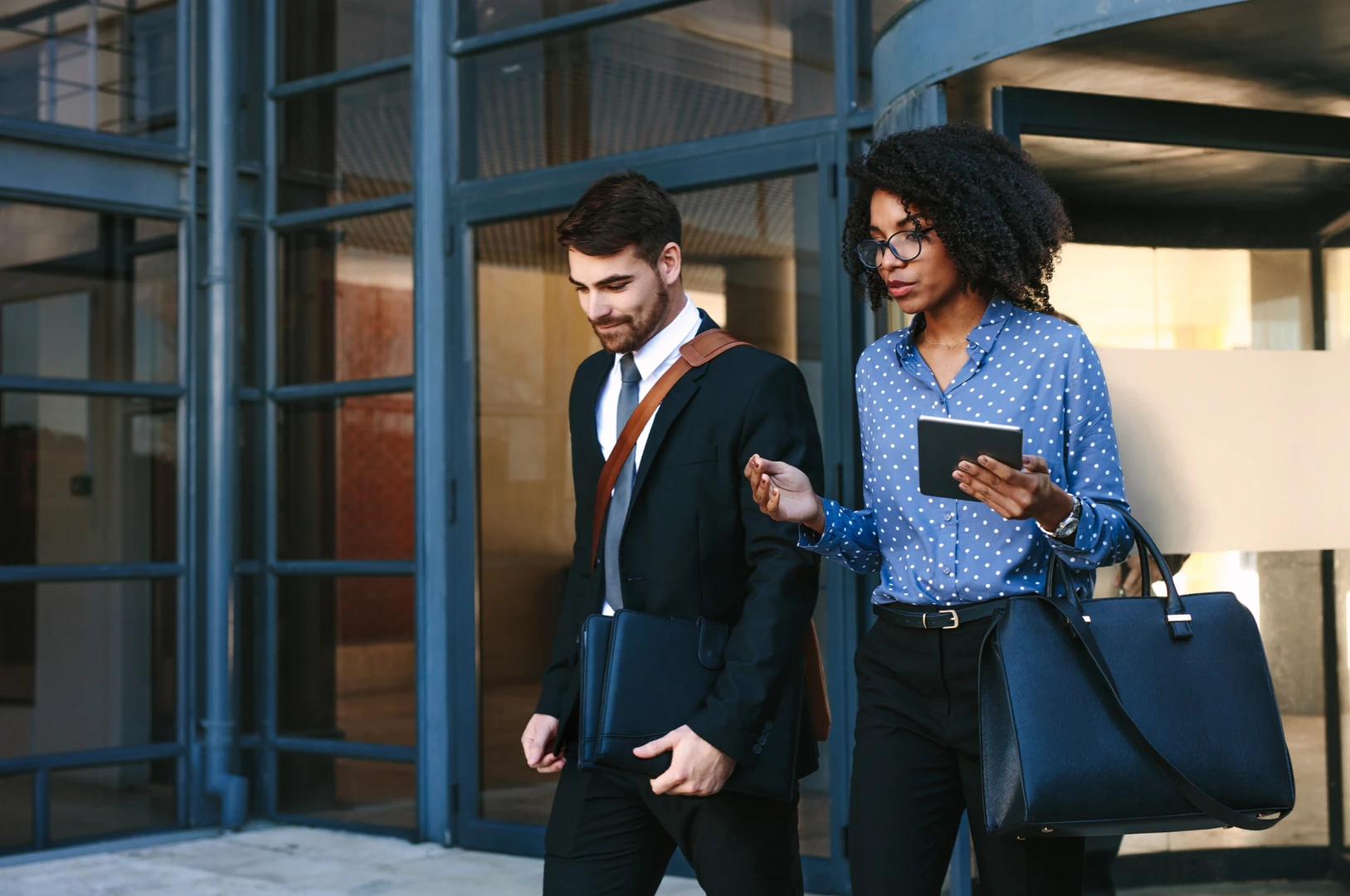 A man in a black suit with a brown shoulder strap and a black portfolio walks alongside a woman in a blue polka dot blouse holding a small tablet and a large black handbag outside a modern glass building.
