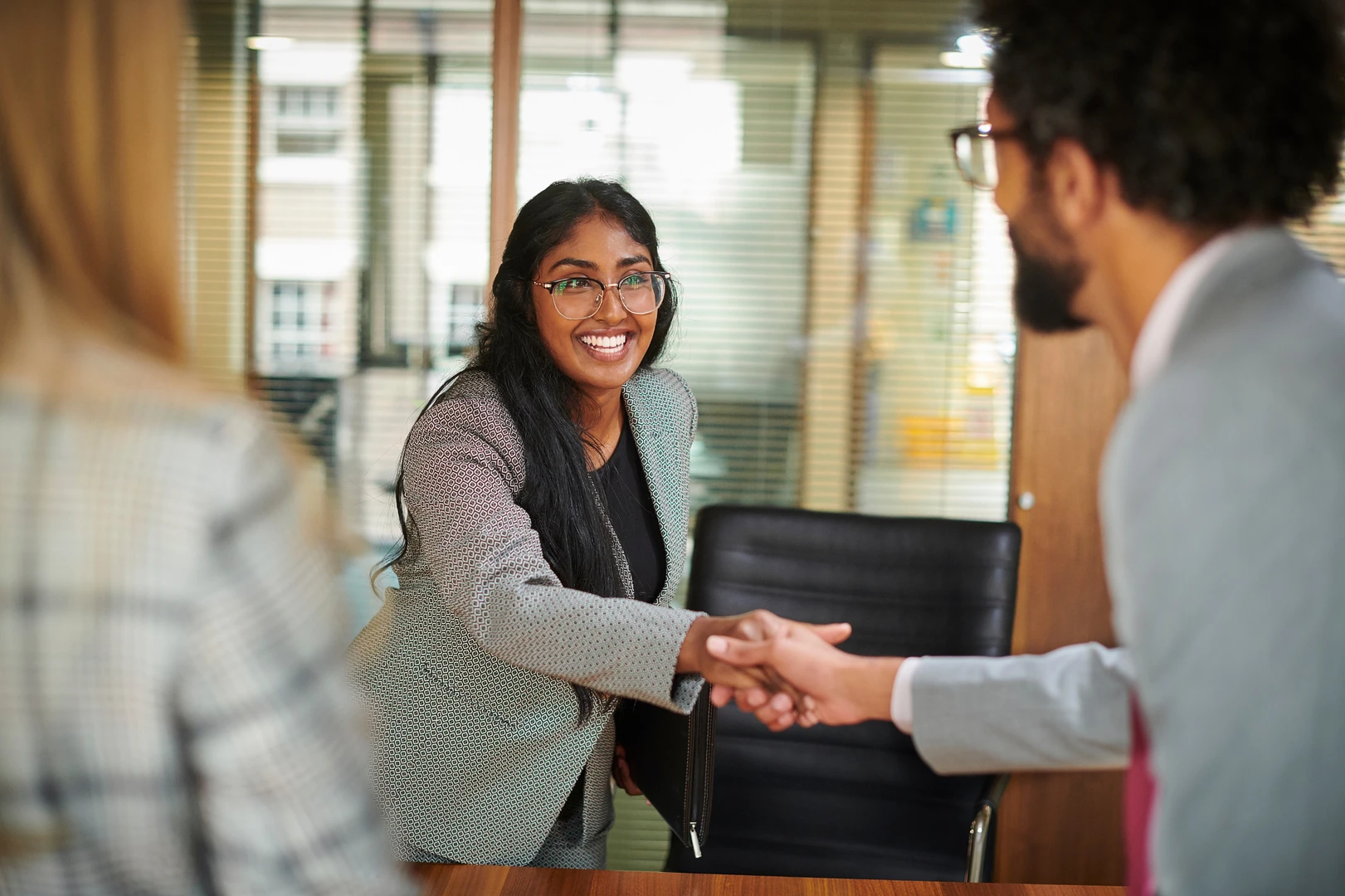 Smiling woman wearing glasses and a patterned blazer shaking hands with a man in a light gray suit across a wooden table in a bright office with glass partitions.