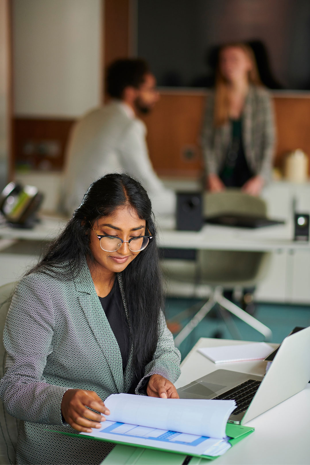 Woman with long black hair and glasses wearing a patterned blazer, sitting at a desk, reviewing a document in a green folder, with an open laptop nearby and two blurred people talking in the background.
