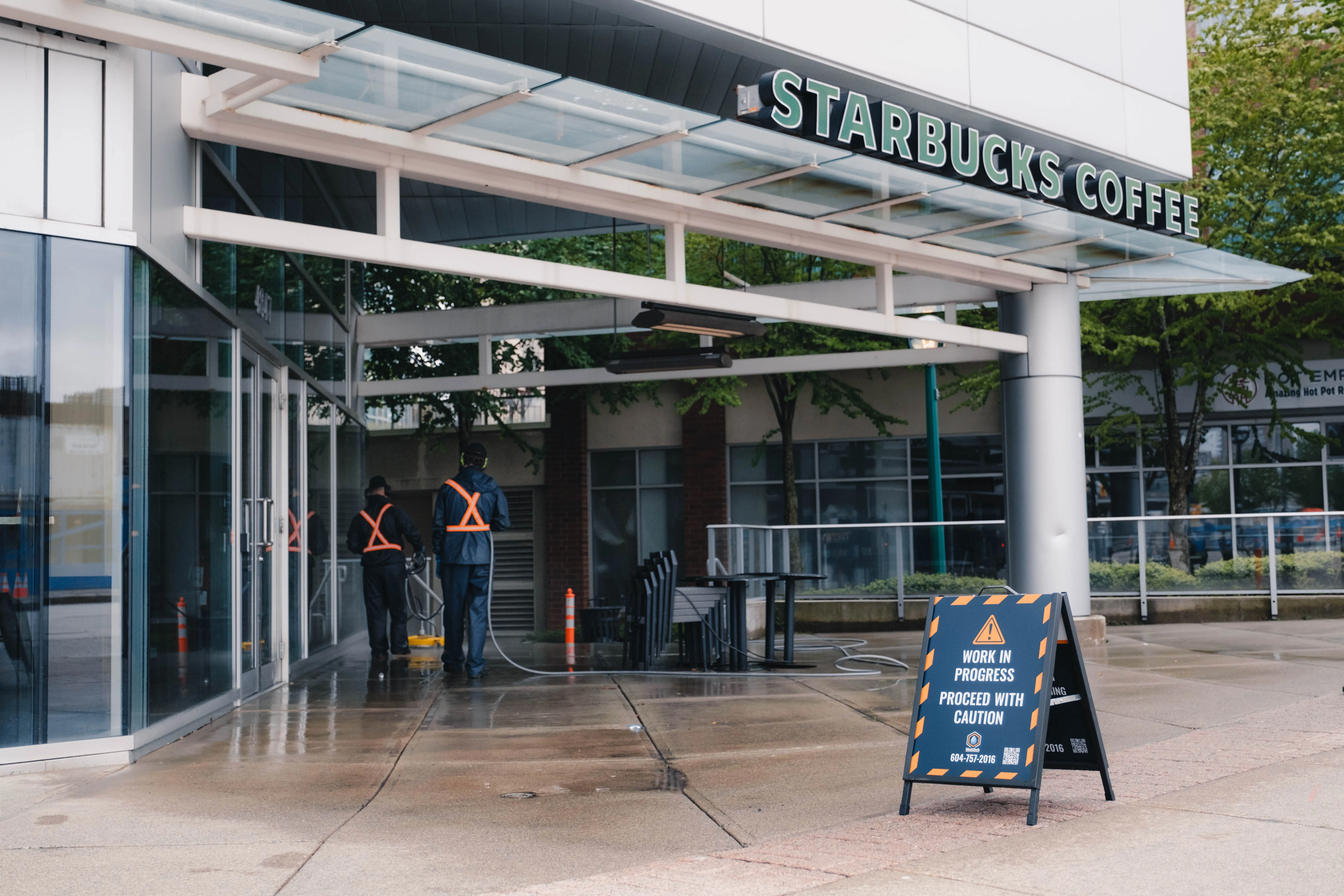 Two WashTech workers in reflective safety vests cleaning a wet sidewalk outside a Starbucks Coffee in Vancouver with a caution sign nearby.