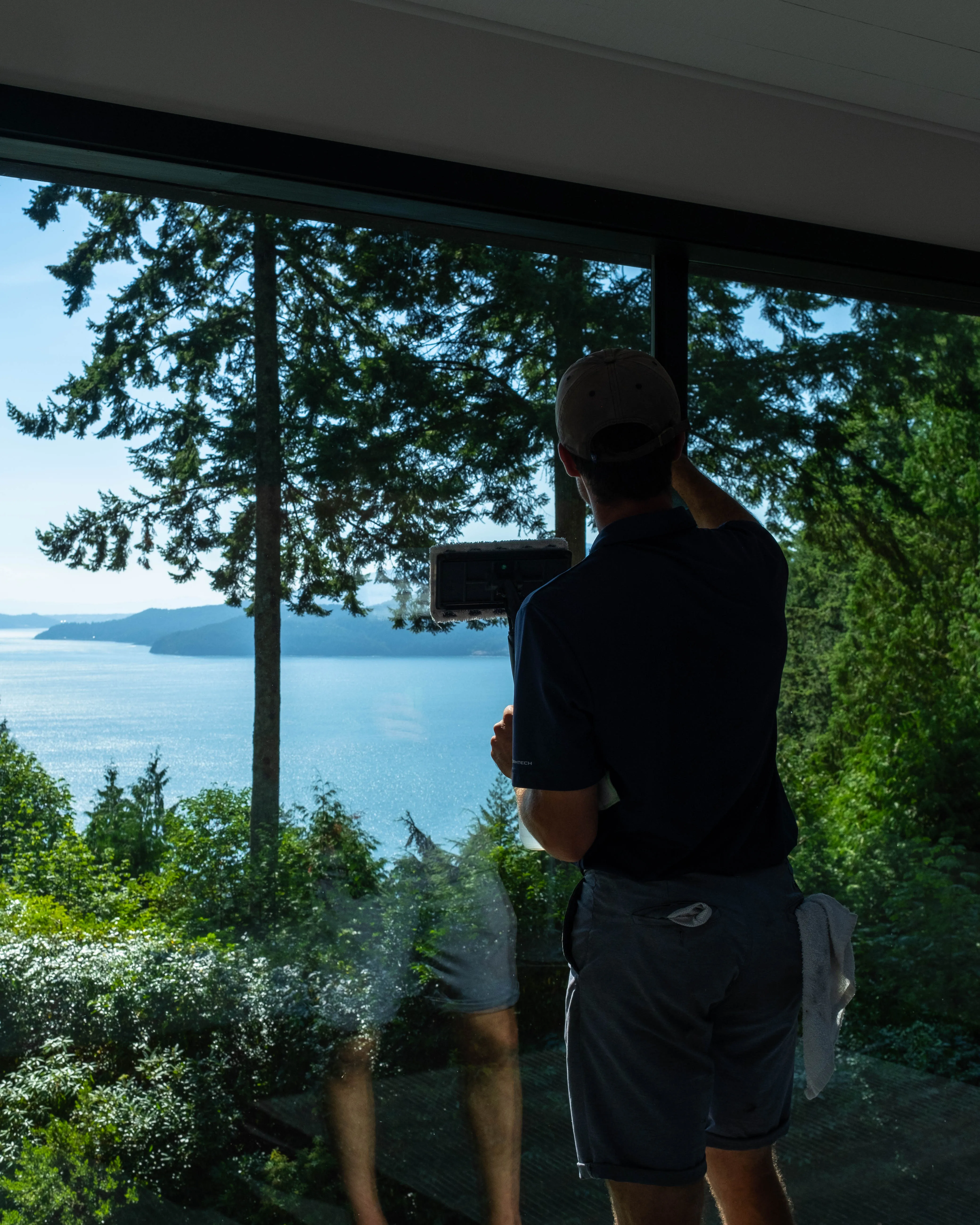 Man cleaning large window inside house with scenic lake and forest view outside.
