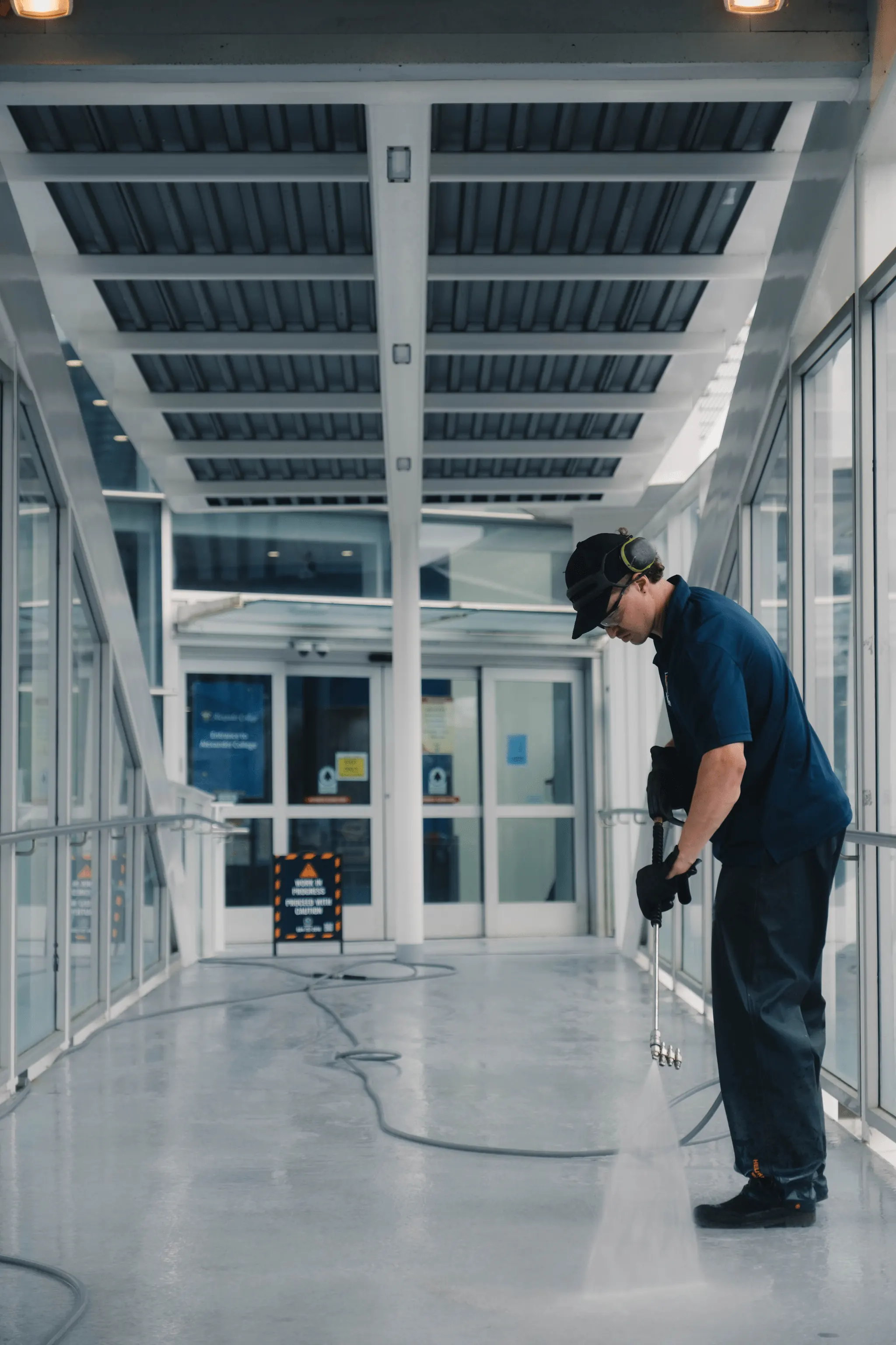 Professional pressure washer cleaning commercial indoor walkway with safety gear and high-pressure water system.