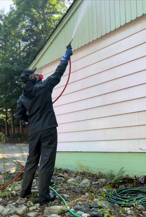 Technician performing soft wash exterior house cleaning with protective gear and pressure wand.