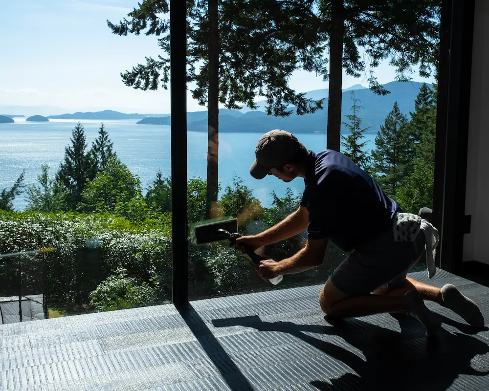 Technician performing interior window cleaning in Lions Bay home with scenic ocean and forest view in the background.