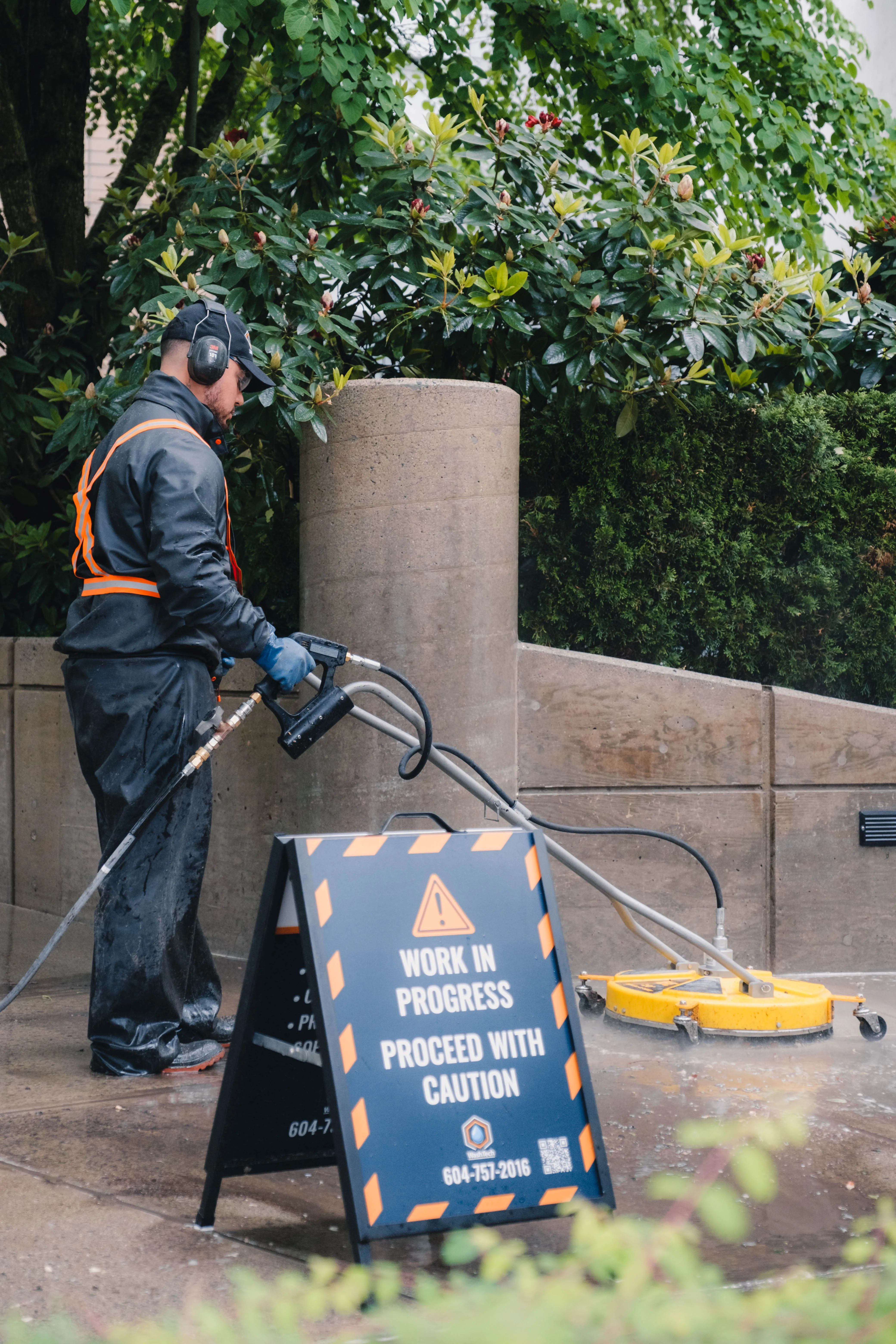 WashTech worker in waterproof clothing power washing a concrete surface with a caution sign reading 'Work in Progress Proceed with Caution' in front.