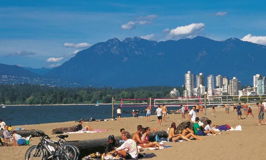 People relaxing and playing volleyball on a sandy beach with bicycles in the foreground, city skyline, and mountains in the background under a blue sky.