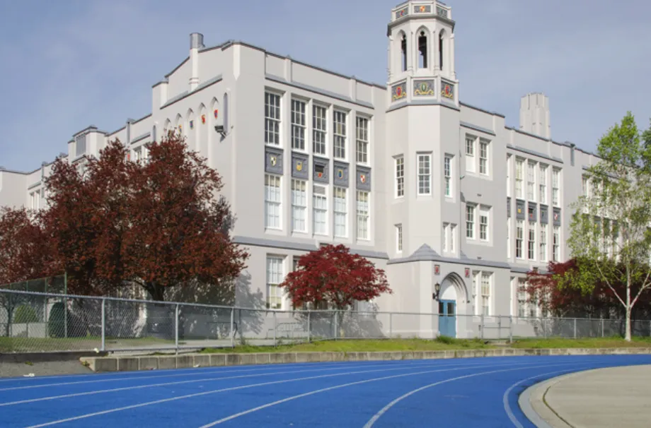 Historic white school building with a tower next to a blue running track and trees with red and green leaves.