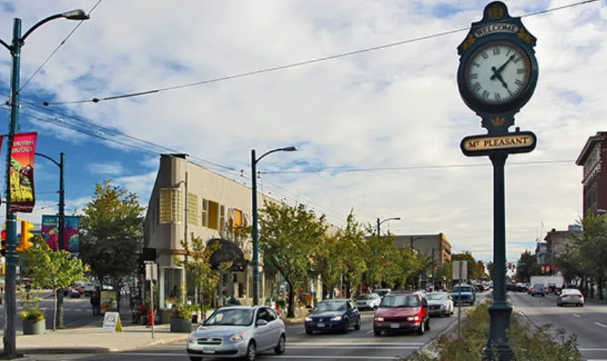 Street view of Mt Pleasant neighborhood featuring a vintage clock showing 10:47, cars on the road, trees, and buildings under a partly cloudy sky.