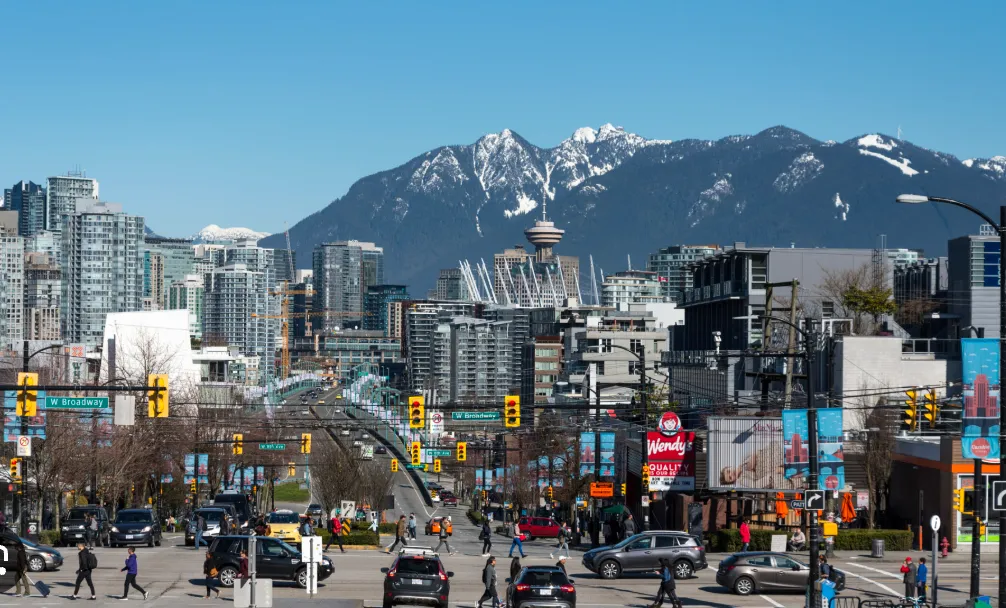 Busy urban street scene with pedestrians crossing, cars waiting at traffic lights, high-rise buildings, and snow-capped mountains under a clear blue sky.