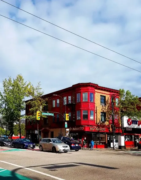 Street corner with a red and beige three-story building housing a restaurant named Red Burrito, cars waiting at a traffic light, and trees on a partly cloudy day.