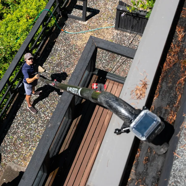Man on ground holding an extended pole with a camera mounted on a rooftop edge.