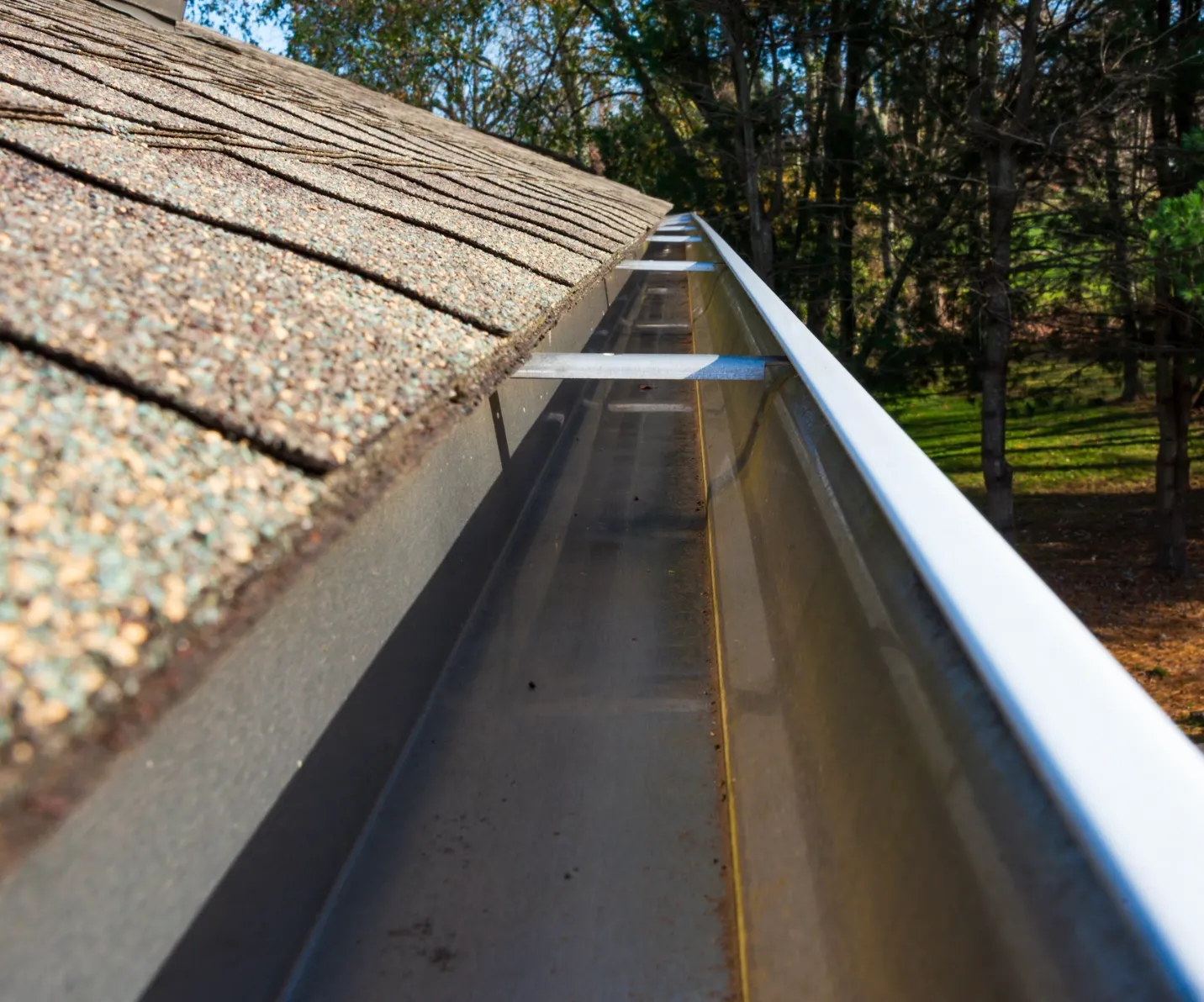 Empty metal rain gutter attached to roof with asphalt shingles beside a wooded area.
