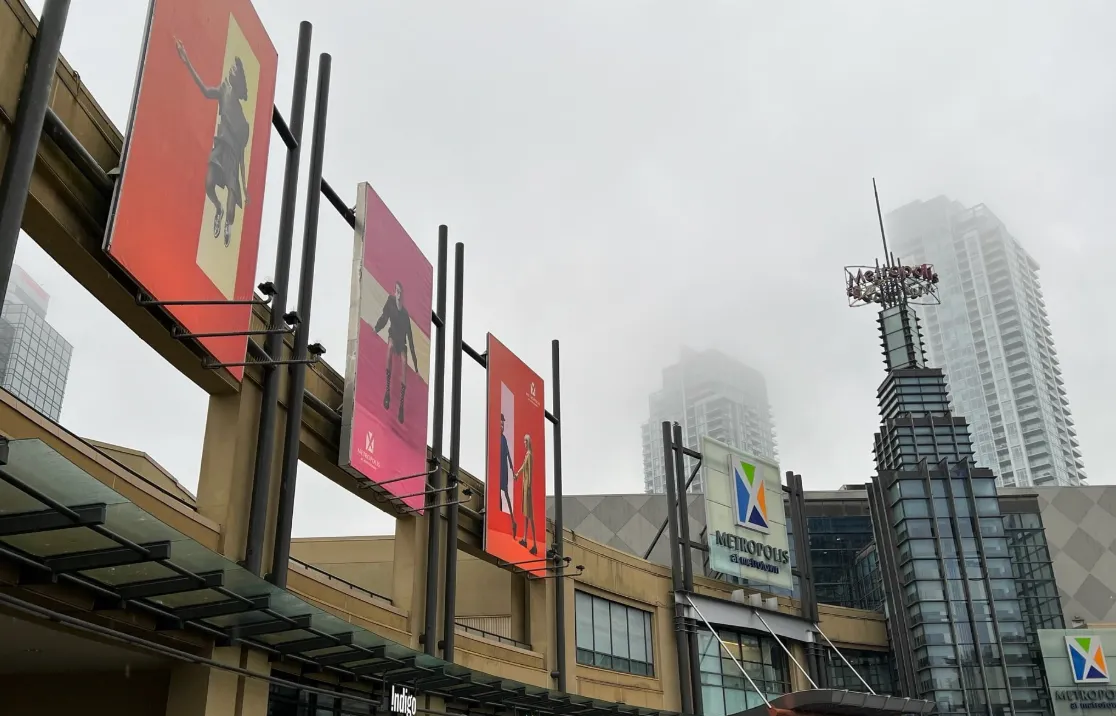 Exterior of Metropolis at Metrotown shopping center with colorful banners and fog-covered high-rise buildings in the background.