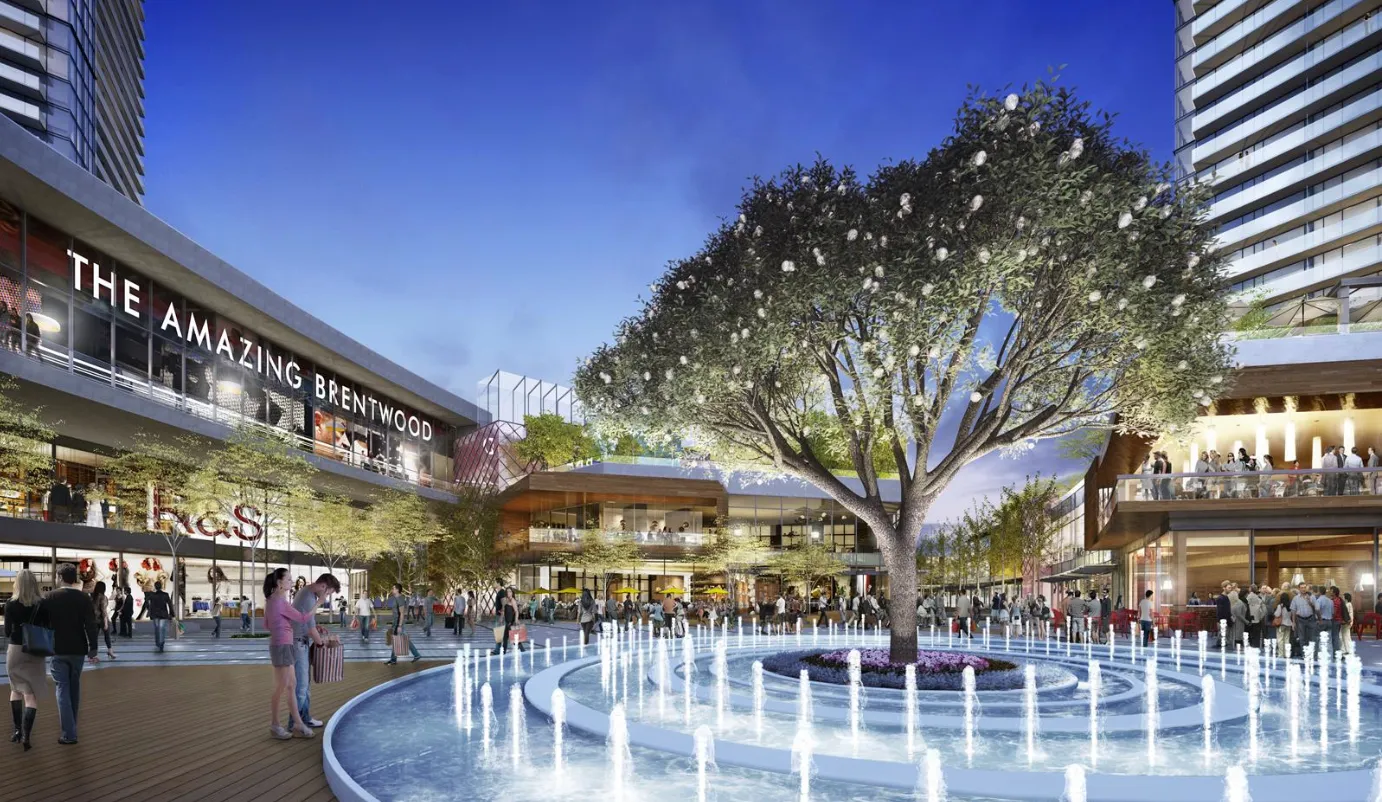 Outdoor plaza at The Amazing Brentwood featuring a large tree with white flowers centered in a modern fountain surrounded by people and retail buildings at dusk.