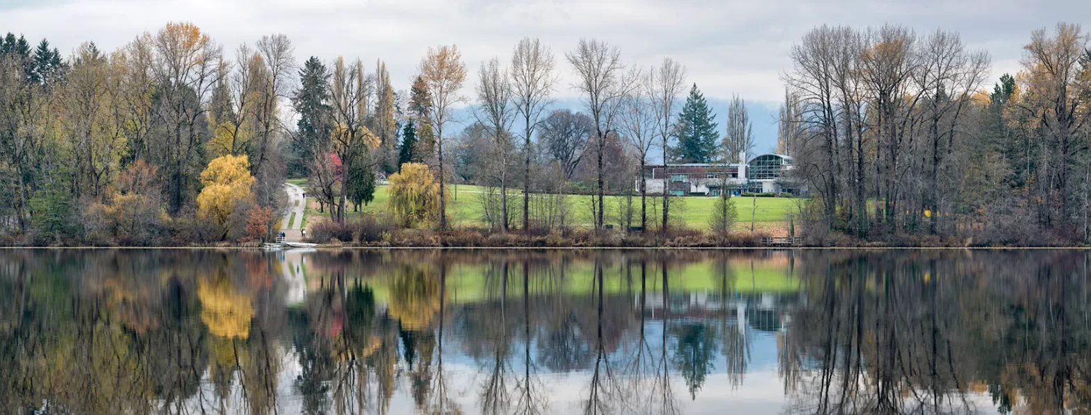 Calm lake reflecting autumn trees and a modern building on a grassy hill under a cloudy sky.