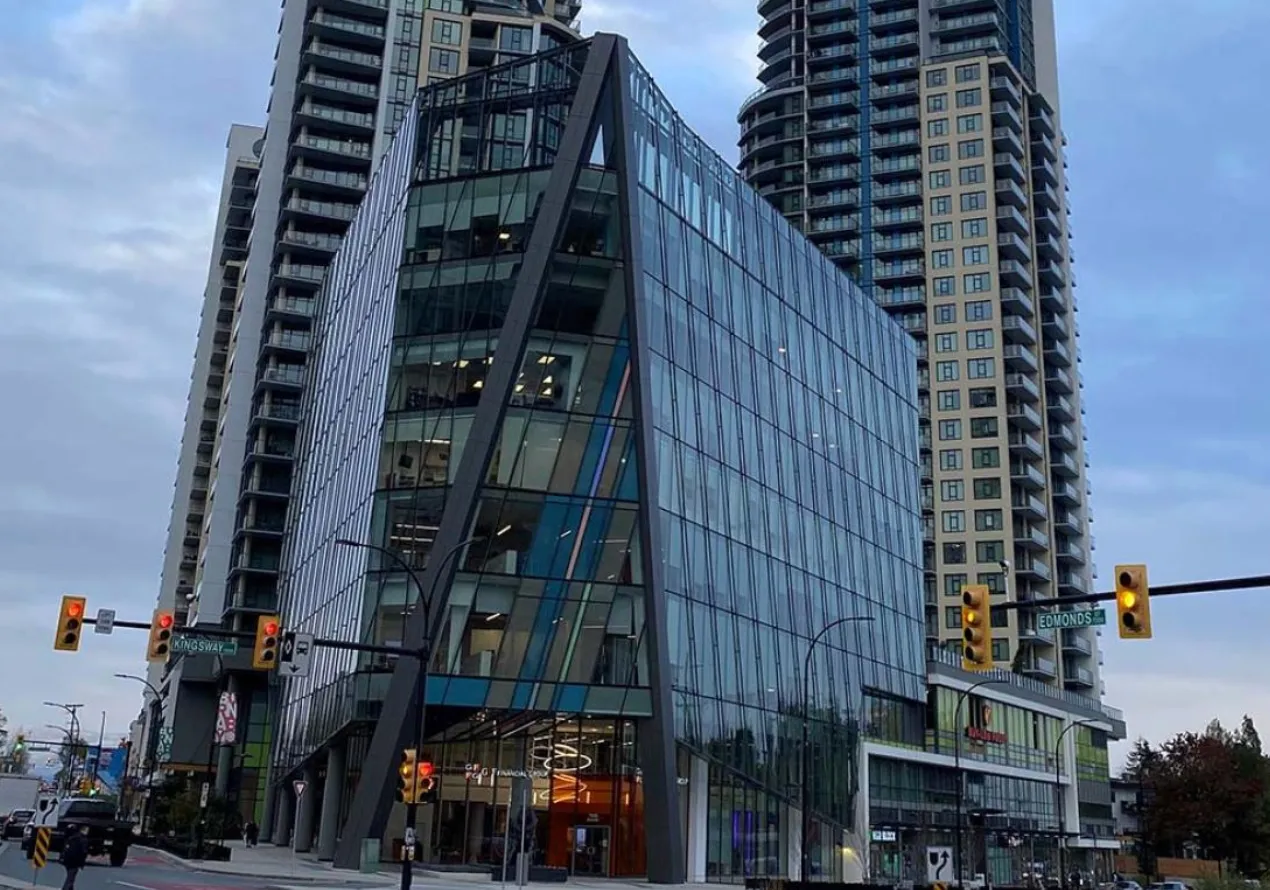 Modern glass office building at an urban intersection with traffic lights showing red and yellow, surrounded by tall residential towers under a cloudy sky.