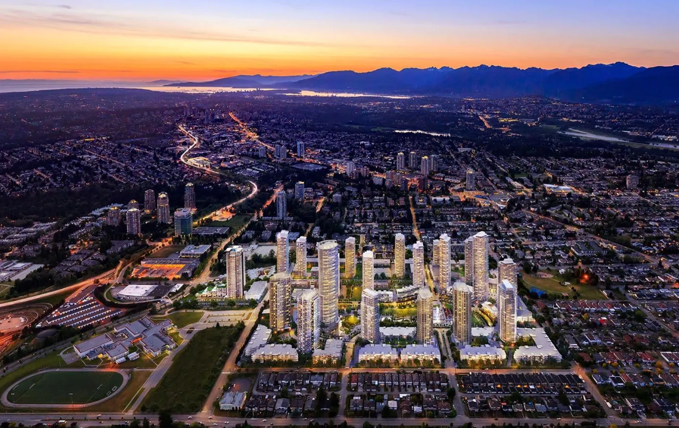Aerial view of a city skyline at sunset, featuring illuminated high-rise buildings and residential areas with mountains in the background.