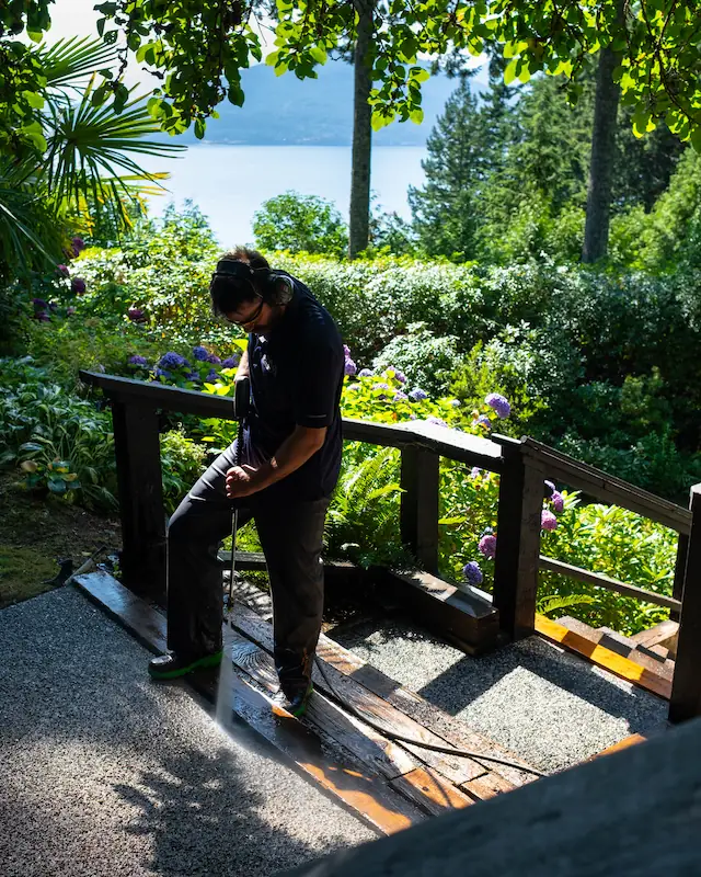 Residential pressure washing in the Vancouver area, with a technician cleaning an outdoor walkway in Lions Bay surrounded by trees, plants, and ocean views.