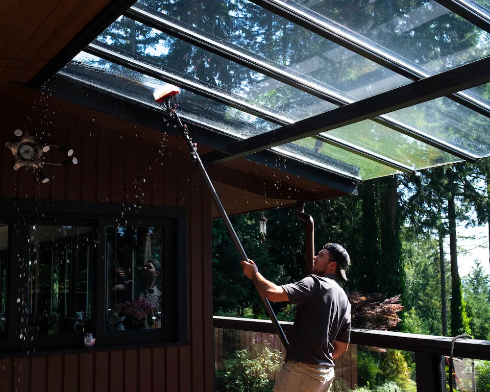 Professional cleaner washing underside of glass awning with extension brush at residential property in Lions Bay.