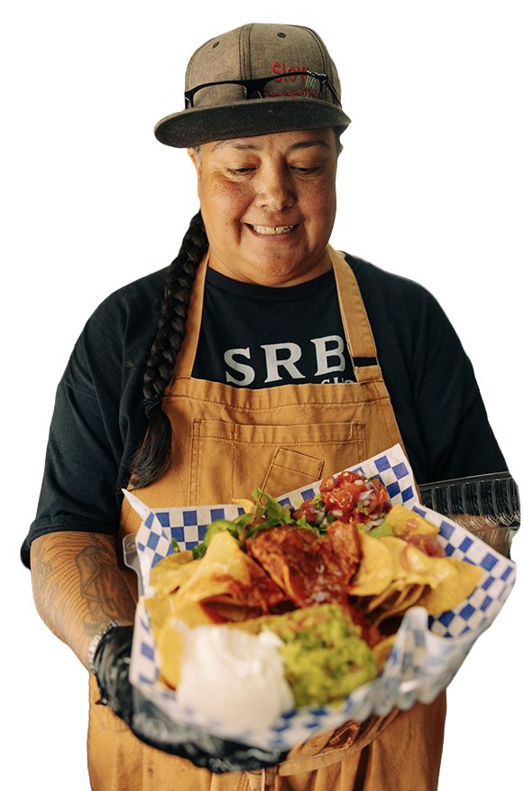 Person wearing an apron and cap holding a tray of loaded nachos with guacamole, sour cream, and salsa in a kitchen.