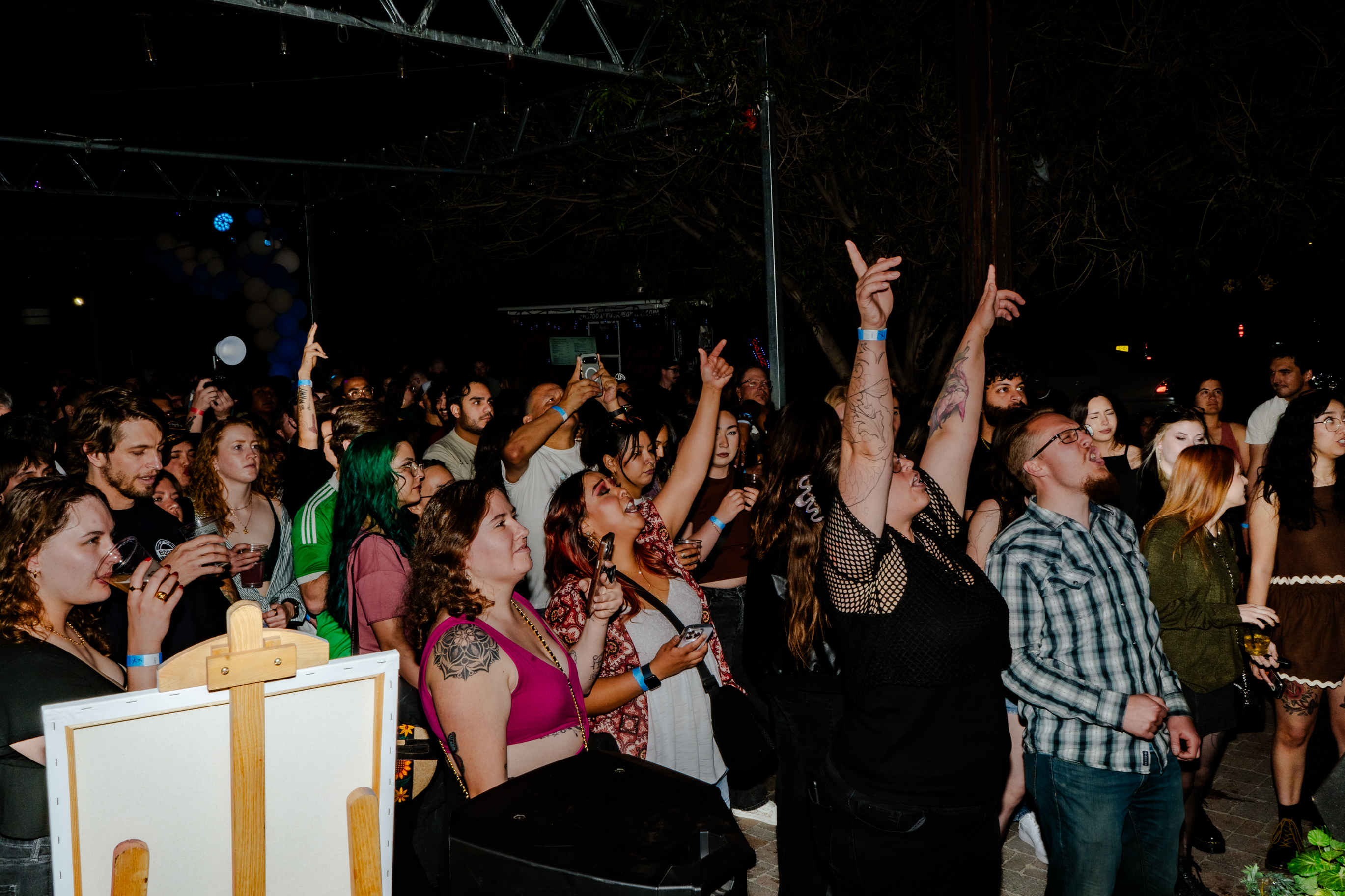 Crowd of people enjoying an outdoor nighttime event, some raising their hands and others holding drinks.