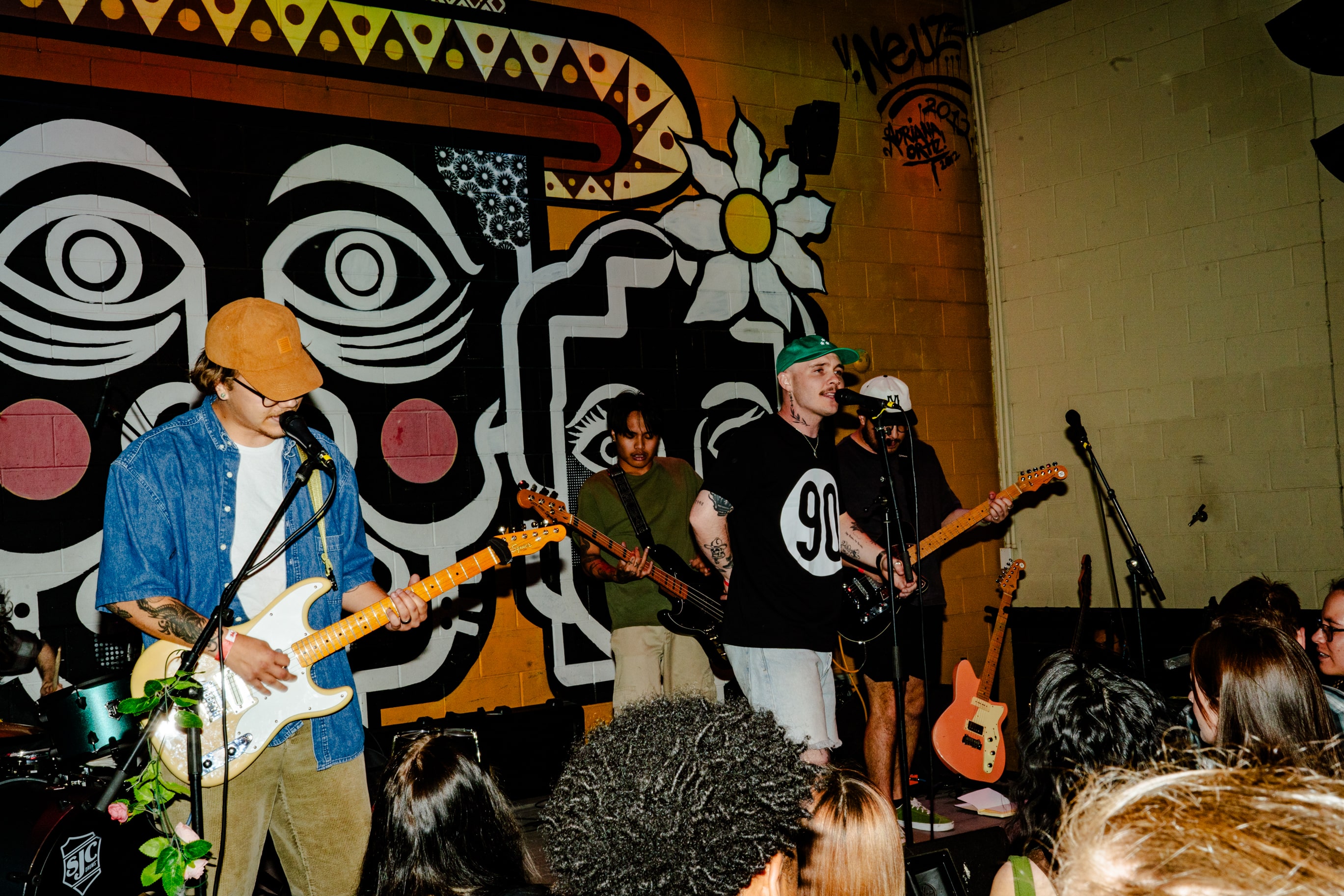 Band performing live indoors in front of a colorful mural with an abstract face and flower design, with audience members watching.