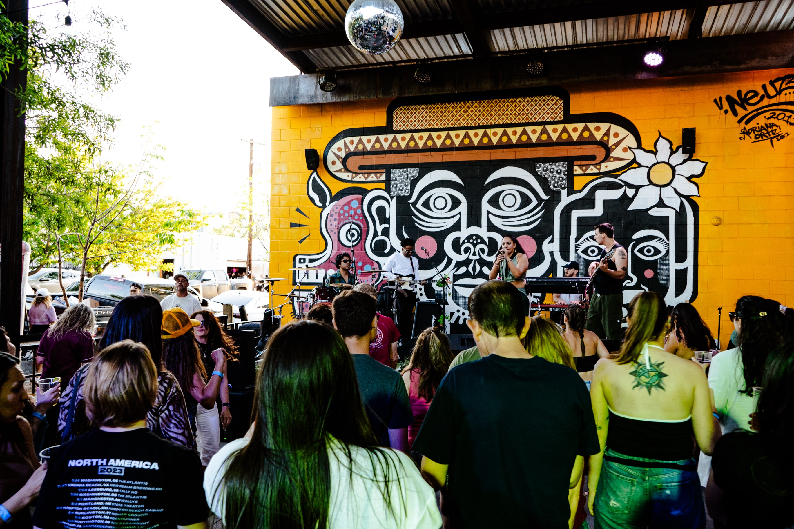 Crowd watching a live band perform in front of a large colorful mural with faces wearing a hat and flower on a yellow wall.