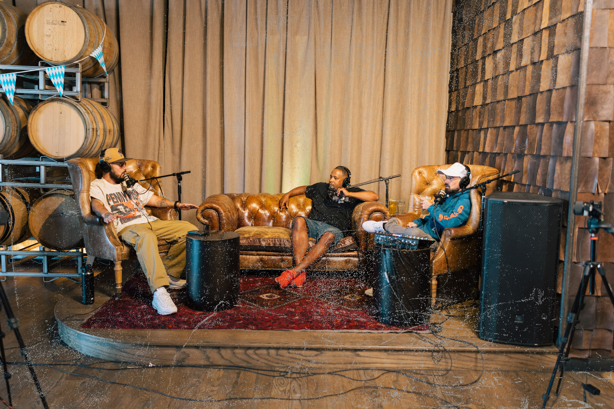 Three men wearing headphones and speaking into microphones seated on leather chairs and a leather sofa in a podcast recording lounge with wooden barrels and rustic decor.