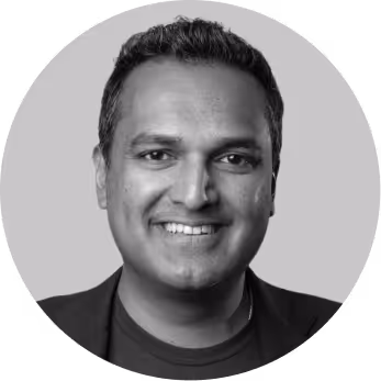 Smiling man with short dark hair wearing a dark jacket and shirt, against a plain background.