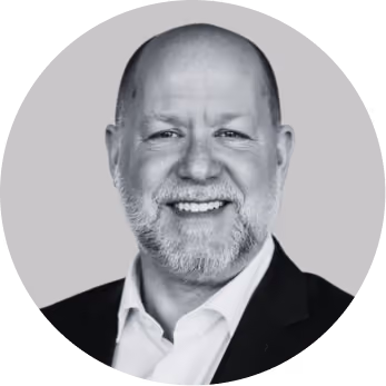 Smiling bald man with beard wearing a white shirt and dark jacket against a gray background.