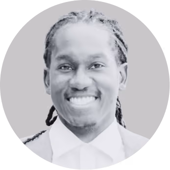 Smiling man with braided hair wearing a suit and white shirt on a gray background.